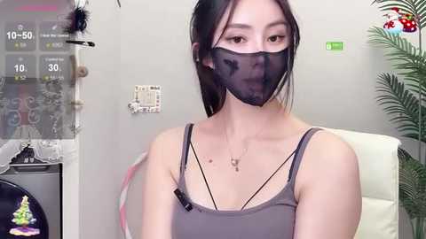 Media: Video of an East Asian woman with light skin and black hair, wearing a black face mask, gray tank top, and multiple thin black necklaces, indoors, near a Christmas tree.