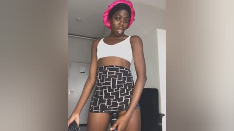 Media: Video of a young Black woman with a muscular build, wearing a white crop top and black patterned skirt, bright pink headband, indoors with a white door and chair in the background.