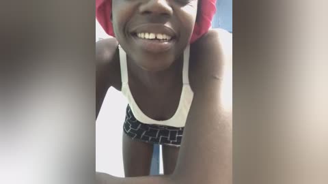 Media: Video of a joyful, dark-skinned woman with a wide smile, wearing a white tank top and black shorts with a geometric pattern. She leans forward, her face filling the frame.