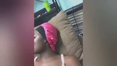 Media: Video of a young Black woman with a bright pink headscarf, lying on a beige couch in a casual living room with a turquoise wall and a green plastic cup.