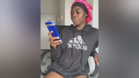 Media: Video of a dark-skinned woman with a pink towel on her head, wearing a black Adidas hoodie and holding a blue water bottle, seated in an office chair against a neutral background.