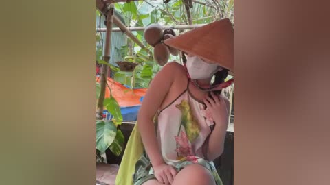 Media: Video of a woman in a green and yellow floral dress, wearing a conical hat, holding a bowl, seated among lush tropical plants.