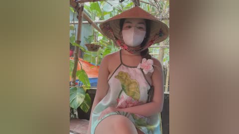 Media: Video of an Asian woman wearing a conical hat, mask, and floral dress, sitting in a lush, tropical garden with hanging plants.