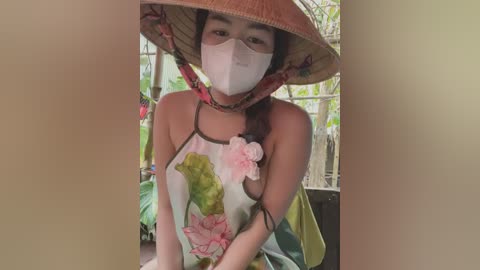 Media: Video of an Asian woman with light skin, wearing a white face mask, floral dress, and conical hat, sitting outdoors amidst greenery and a wooden structure.