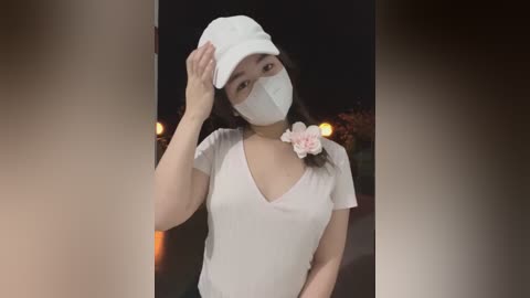 Media: Video of an Asian woman wearing a white face mask, cap, and t-shirt, adjusting her cap, with blurred, dark background.