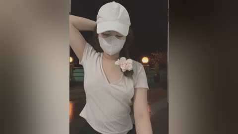 Media: Video of a young East Asian woman in a white cap, face mask, and light pink shirt with a rose brooch, standing in a dimly lit outdoor setting at night.