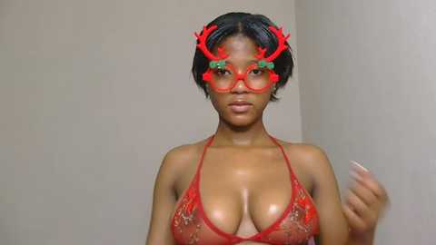 Video of a young Black woman with medium skin tone, short black hair, wearing a red lace bra, red antler headband, and red-tinted glasses, indoors against a beige wall.