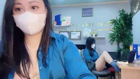 Media: Video of a woman in a blue shirt, face mask, and long dark hair, sitting in a modern office with plants and framed certificates on the wall.