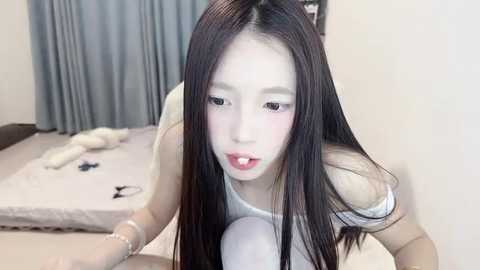 Media: A video of an Asian woman with long black hair, wearing a white off-shoulder top, kneeling on a bed in a minimalist room. She has a white substance smeared on her face.