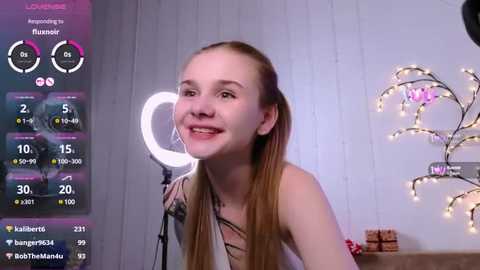 Media: Video of a smiling young woman with long blonde hair, wearing a sleeveless top, standing in front of a white wall with a heart-shaped string of lights and a glowing ring light.