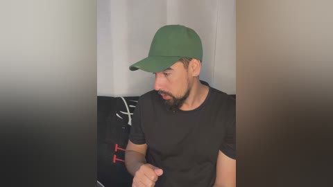 Media: Video of a light-skinned man with a beard and medium-length hair, wearing a green cap and black t-shirt, sitting on a black couch.