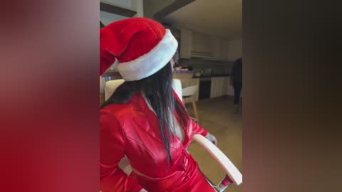 Media: Video of a woman in a red velvet Santa hat and matching dress, seated in a modern kitchen with white cabinets and beige countertops.