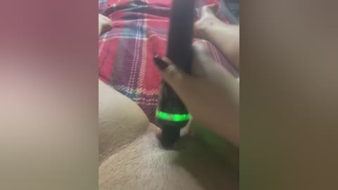 Media: A video of a person with a light skin tone using a black sex toy with a green light on their exposed genitals, wearing a red plaid shirt.