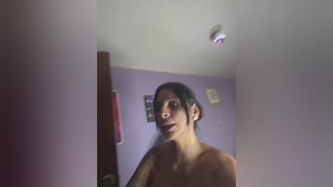 A blurry video of a topless woman with tattoos, medium build, and short dark hair, standing in a dimly lit room with lavender walls and a small, red light fixture.