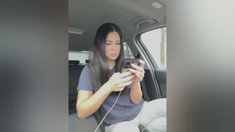 Media: Video of a Latina woman with long black hair in a gray t-shirt, sitting in a car, texting on her phone, wearing a gray sweater.