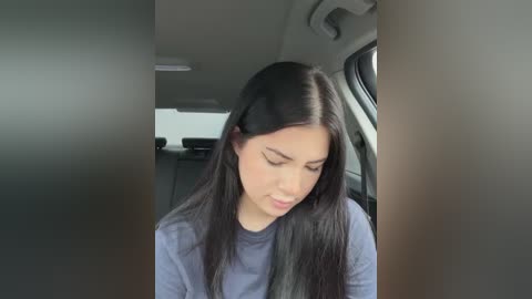 Media: Video of a young Asian woman with long black hair, wearing a grey shirt, seated in a car with grey upholstery, looking down.