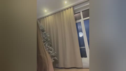 Video of a cozy, warmly lit room with beige curtains, a snowy Christmas tree, and a lit window showcasing a moonlit night.