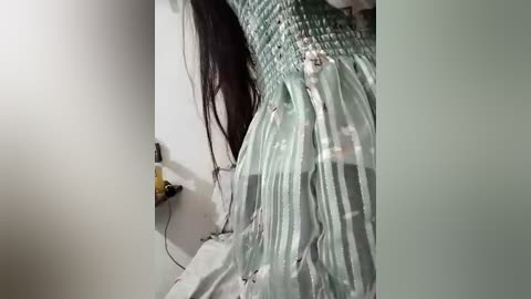 Media: Video of a woman with long, dark hair, wearing a flowing, green and white striped dress, standing in a dimly lit room with a white wall and a yellow power outlet visible on the left.