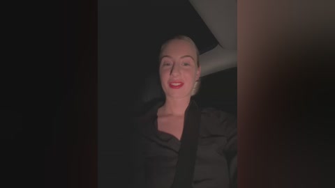 Media: Video of a smiling woman with light skin, short blonde hair, red lipstick, wearing a black shirt, seated in a dark car interior.