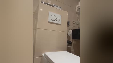 Media: A video of a small, beige-tiled bathroom with a white wall-mounted refrigerator above a white toilet. The refrigerator has a control panel with two buttons. A white phone and a towel holder are visible in the background.