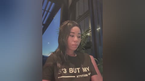 Media: Video of a young woman with long dark hair wearing a black T-shirt with the phrase \"I'm 13 but my attitude is adult\" in white letters. Background shows a dark room with a window and green plants.