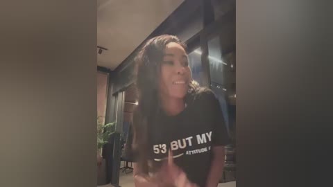 Media: Video of a smiling young woman with long, wavy hair wearing a black T-shirt with \"53 BUT MY ATTITUDE IS\" in white text. She stands in a dimly lit, modern indoor space with a potted plant in the background.