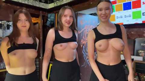 Media: Video of three topless Caucasian women wearing black sports bras and high-waisted black leggings in a dimly lit bar. The background features a colorful neon sign and a shelf with bottles.