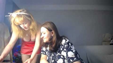 Media: Video of two women in a bedroom: one blonde in a red top, the other brunette in a floral shirt. Background features a white bed with stuffed toys.