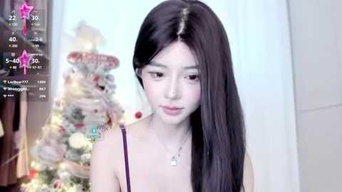 Media: Video of a young East Asian woman with long, straight black hair, fair skin, and delicate facial features, wearing a purple spaghetti-strap top, standing in front of a decorated Christmas tree.