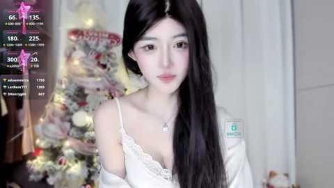 Media: A video of a young East Asian woman with long black hair, wearing a white off-shoulder top, standing in a softly lit room with a decorated Christmas tree in the background.