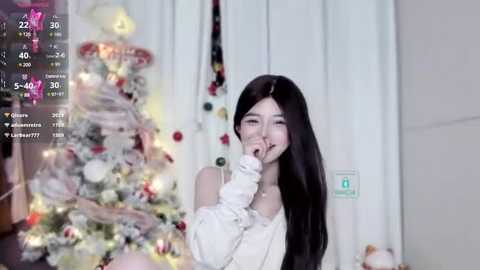 Media: A video of a young Asian woman with long black hair, wearing a white top and long gloves, playfully covering her mouth while standing in front of a decorated Christmas tree in a softly lit room.