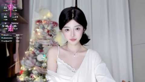 Media: Video of an Asian woman with fair skin, dark hair, and a white off-shoulder top, standing in front of a festive Christmas tree adorned with lights and ornaments.