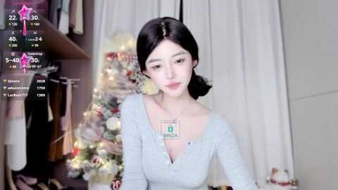 Media: Video of a young East Asian woman with long black hair in a side braid, wearing a light gray V-neck sweater, sitting in a cozy, dimly lit room with a decorated Christmas tree and a white curtain in the background.