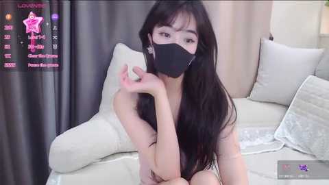 Media: A video of an Asian woman with long black hair and a black face mask, sitting on a bed with white pillows and blankets.