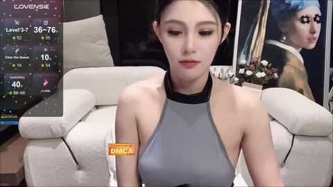 A video of a young East Asian woman with fair skin, dark hair, and a slender physique, wearing a gray halter top, seated in a modern living room with a white couch and a TV displaying a weather forecast.