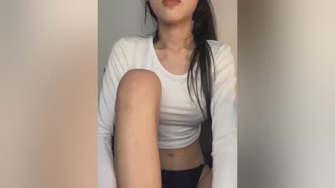 Media: Video of an East Asian woman with long black hair, fair skin, and medium-sized breasts, wearing a white long-sleeve crop top and black panties. She sits with her legs bent, one leg raised, against a plain beige wall.