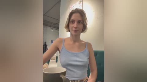 Media: Video of a young woman with short blonde hair, wearing a light blue tank top, sitting in a teal booth in a modern, dimly-lit restaurant.