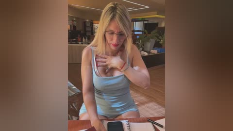 Media: Video of a blonde woman with glasses and a blue tank top, sitting at a wooden table in a cozy, modern living room.