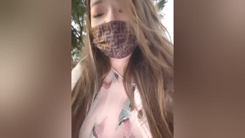 Media: A video of a woman with long brown hair, wearing a floral-patterned shirt and a face mask with a leopard print, taken outdoors with blurred greenery in the background.