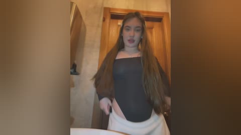 Media: Video of a young woman with long, straight brown hair, wearing a black, long-sleeve bodysuit and white skirt, standing in a beige-tiled bathroom with wooden door in the background.