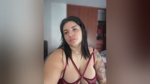 Media: Video of a plus-size woman with dark hair, wearing a revealing red strappy bra, showcasing tattoos on her left arm, standing in a bedroom with wooden furniture and a bed in the background.