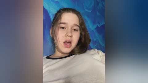 Media: Video of a young Caucasian girl with long brown hair, wearing a white long-sleeved shirt, set against a blue, abstract background. She has a neutral expression with slightly parted lips.