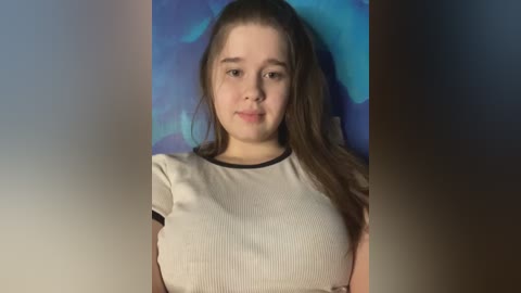 Media: Video of a young Caucasian girl with fair skin, long brown hair, and a neutral expression, wearing a white ribbed top. Background features a blue abstract mural.