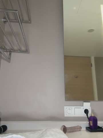 Media: Video of a modern, minimalist hotel room with a large, light grey wall, a mirrored closet door, a metal shelf, and a white outlet.