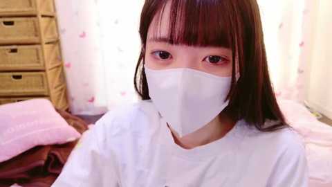 Media: Video of an East Asian woman with straight, brown hair and bangs, wearing a white surgical mask and white t-shirt, sitting on a bed with pink sheets, next to a wicker basket, in a softly lit room with floral curtains.