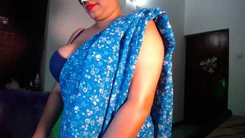 Media: Video of a woman with light brown skin, wearing a blue floral sari, red lipstick, and glasses, standing indoors near a dark wooden door.