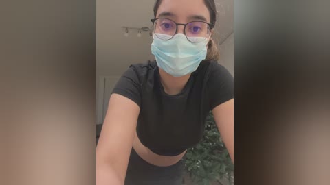 Media: A video of a young woman with light skin, brown hair, and glasses, wearing a blue face mask and a black crop top, leaning forward in an indoor setting with blurred background.