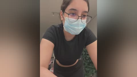 Media: Video of a young woman with fair skin, brown hair, and round glasses, wearing a blue surgical mask and black short-sleeve crop top. Background is blurred, featuring green plants.