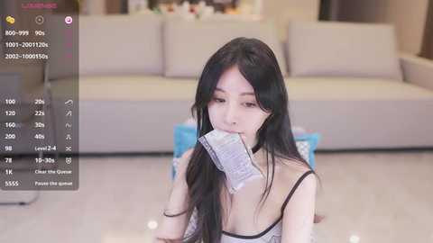 Media: A video of an Asian woman with long black hair, wearing a revealing black lace bra, eating a packet of chips on a beige carpet in a modern living room.