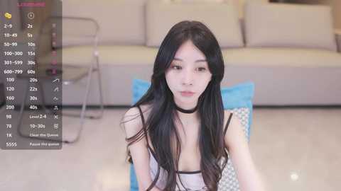 Media: Video of a young East Asian woman with long black hair, fair skin, and a slender physique, wearing a black choker and a sheer, white top. She sits on a blue cushion in a modern living room with beige furniture.